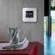 Digitalthermostat von Theben an grauer Wand, mit Bildschirm zur Temperaturanzeige und Tasten zur Einstellung, daneben ein Glas mit Markern und Zeichnungen auf einem roten Tisch.